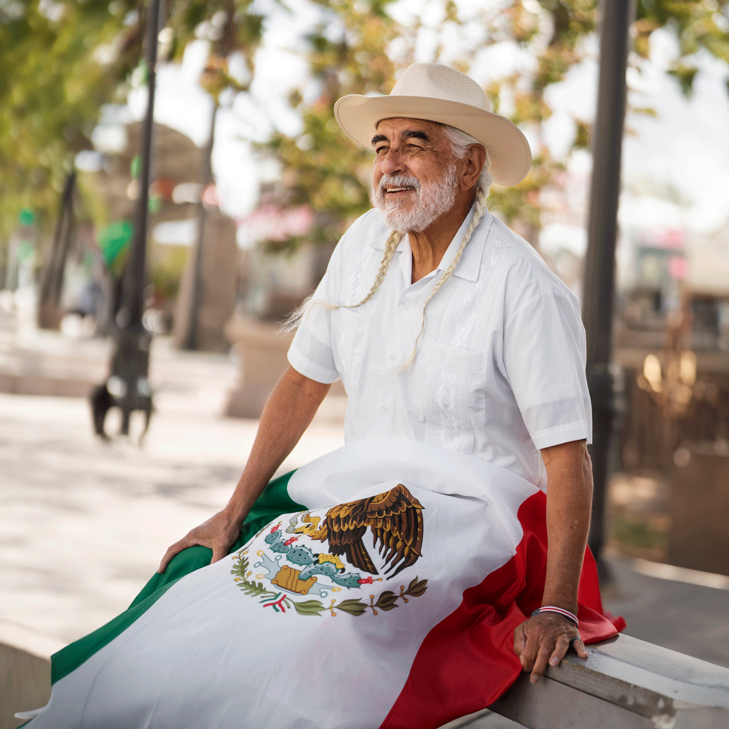 Hombre mayor con el pelo blanco y trenzas. Tiene en sus piernas una bandera de México.